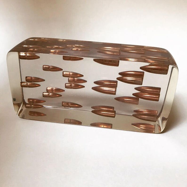 May include: A clear rectangular block filled with copper-colored bullet casings. The casings are arranged in a random pattern and are visible from all sides of the block.