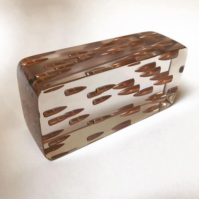 May include: A clear rectangular block of resin with multiple small, brown bullets embedded inside. The bullets are arranged in a random pattern.