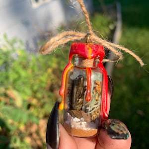 Op de afbeelding: Een klein glazen flesje met een rode en gele waszegel. Het flesje is gevuld met verschillende kristallen, kruiden en andere natuurlijke materialen. Het flesje hangt aan een touwtje.