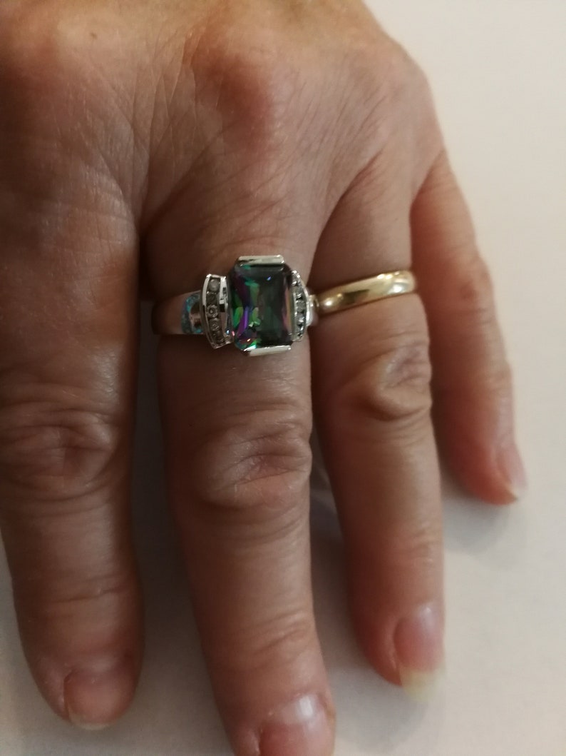 May include: A silver ring with a large, rectangular, iridescent gemstone surrounded by smaller clear gemstones. The ring is on a person's finger.