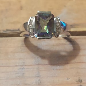 May include: A silver ring with a large, rectangular, iridescent green gemstone surrounded by smaller clear gemstones.