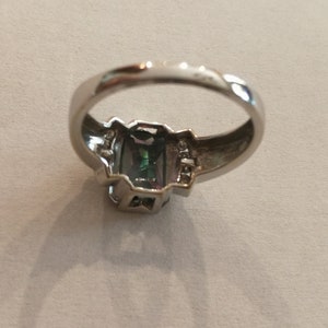 May include: A silver ring with a large, faceted, green and blue gemstone set in a silver bezel. The ring has a simple band and is adorned with smaller clear gemstones on either side of the main stone.