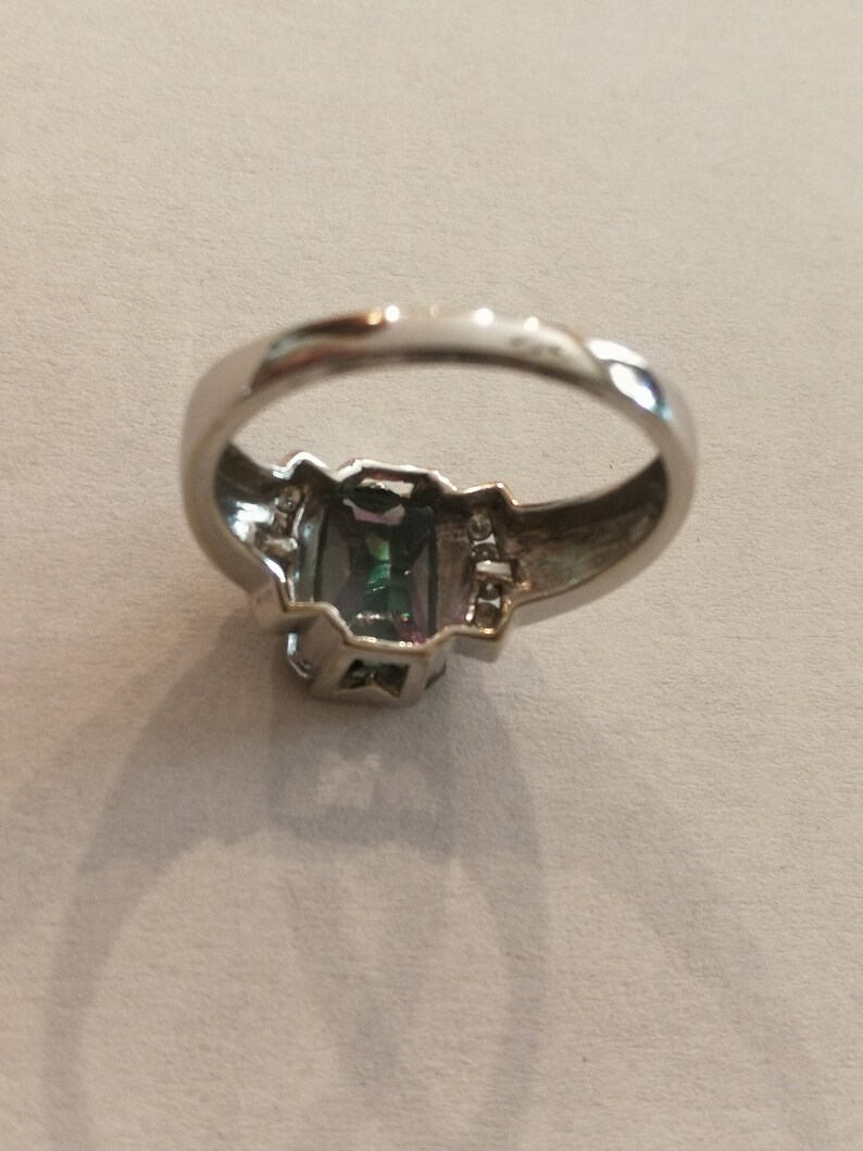May include: A silver ring with a large, faceted, green and blue gemstone set in a silver bezel. The ring has a simple band and is adorned with smaller clear gemstones on either side of the main stone.