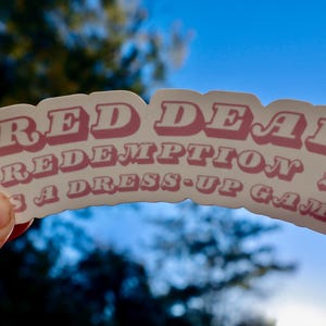 May include: A sticker with the text "RED DEAD REDEMPTION 2 IS A DRESS-UP GAME" in pink letters with a white outline. The sticker is held up against a blue sky and tree background.