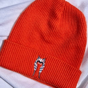 以下が含まれることがあります： リブ編みの鮮やかなオレンジ色のビーニー帽。帽子にはキャラクターの顔の刺繍デザインがあります。帽子は柔らかい素材でできており、肌寒い季節に適しています。冬のアクセサリーです。