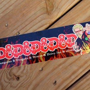 May include: A rectangular sticker with the text "D&D & D&D" in red, with dragon illustrations, and the words "Dungeons & Dragons & Diners & Drive-Ins & Dives" below. The sticker features a cartoon image of a man with sunglasses and a flame background.