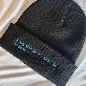 Puede incluir: Gorro de punto acanalado negro con el texto bordado "Hace mucho tiempo, en una galaxia muy, muy lejana..." en azul claro. El gorro está doblado en el borde.