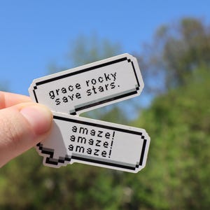 Puede incluir: Dos pegatinas blancas con bordes pixelados negros se sostienen contra un cielo azul y follaje verde. La pegatina superior dice "grace rocky save stars". La pegatina inferior dice "amaze! amaze! amaze!"