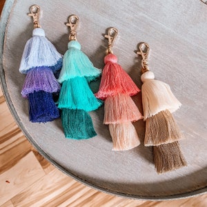 May include: Five colorful tassel keychains with gold metal rings. The tassels are layered in different colors, including blue, purple, green, pink, and beige.
