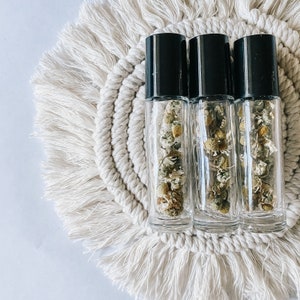 May include: Three clear glass roller bottles with black lids filled with dried chamomile flowers. The bottles are sitting on a white crocheted mat.