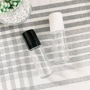 Könnte beinhalten: Zwei transparente Glas-Rollflaschen mit schwarzen und weißen Kappen. Die Flaschen befinden sich auf einem weißen und grauen gestreiften Stoff.