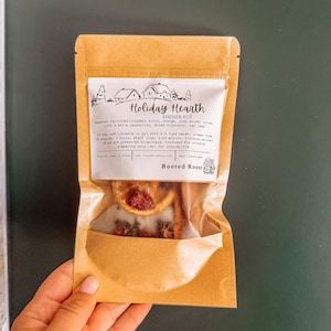 Op de afbeelding: Een bruin papieren zakje met een Holiday Hearth sudderpotmix. Het etiket bevat een lijntekening van huizen en tekst met ingrediënten zoals kaneel, sinaasappel, steranijs, kruidnagel, peperkorrels, veenbessen en laurierblad voor op het fornuis.