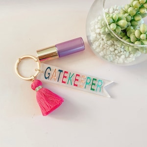 Puede incluir: Un llavero de acrílico transparente con la palabra "GATEKEEPER" escrita en letras de colores. El llavero tiene un borla rosa y un anillo de llave dorado. También se ve una botella morada y dorada en la imagen.