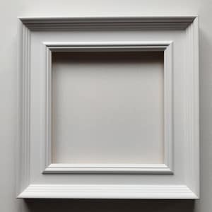 May include: A square, white picture frame with a layered design. The frame has a recessed center and a textured surface. The frame is made of wood and is suitable for displaying artwork or photographs.