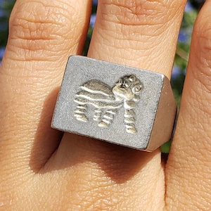 May include: A silver ring with a square-shaped setting featuring a raised design of a stylised animal. The ring is worn on a person's finger.