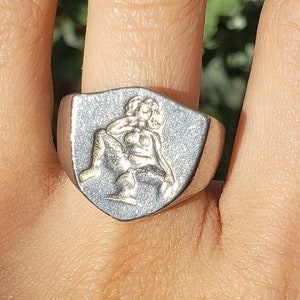 May include: A silver ring with a shield-shaped design featuring a raised image of two figures.