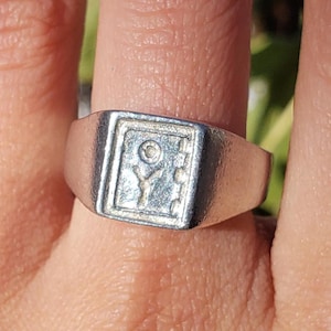 May include: A silver ring with a square-shaped design featuring a stylized flower with a small circle in the center.