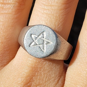 May include: A silver-toned signet ring with a raised, circular face. The face features a five-pointed star design. The ring is worn on a finger, with the background out of focus.