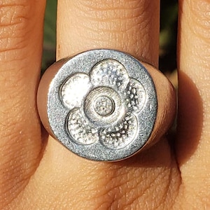 May include: A silver ring with a floral design. The ring features a circular face with a raised flower pattern. The flower has a central circle surrounded by six petals. The ring is worn on a finger.