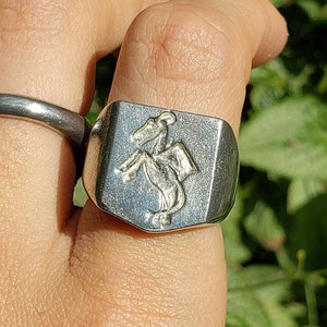 Puede incluir: Un anillo de plata con un diseño en relieve de un caballero montando un caballo. El anillo se lleva en el dedo de una persona.