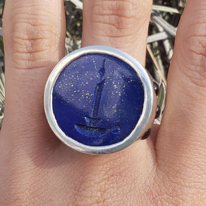 May include: A silver ring with a round blue lapis lazuli stone set in a silver bezel. The stone has a silver design of a candle on it.
