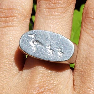 May include: Silver-toned ring with an oval face featuring an engraved design of a duck and three ducklings. The ring is worn on a hand with red nail polish. The ring is a piece of jewelry.