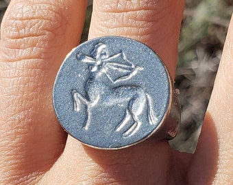 May include: A silver ring featuring a raised design of a centaur holding a bow and arrow. The ring is worn on a person's finger.