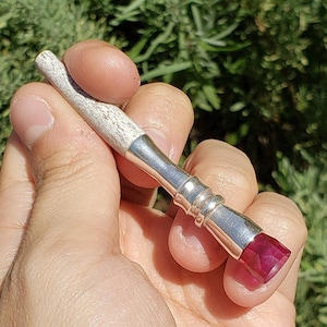 May include: A white and silver pipe with a red gemstone on the end. The pipe is held in a hand.