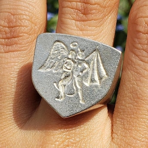 May include: A silver ring with a shield-shaped design featuring an angel and a devil. The ring is worn on a person's finger.