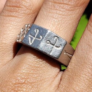 May include: A silver ring with a rectangular band featuring etched symbols. The ring has a textured design on one side and two symbols on the front. The ring is worn on a finger.