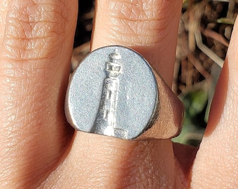 May include: A silver-toned signet ring featuring a circular face with an embossed lighthouse design. The ring has a smooth, polished finish and a wide band that tapers towards the back, showcasing a detailed relief of a lighthouse structure.