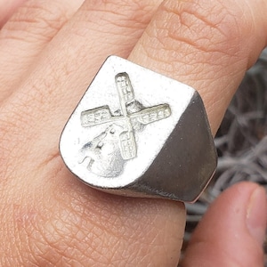May include: A silver ring featuring a windmill design. The ring is worn on a person's finger.
