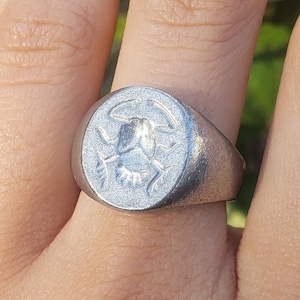 May include: A silver-toned signet ring featuring a circular face with an engraved scarab beetle design. The ring has a matte, textured finish and a wide band that tapers toward the back, showcasing a detailed insect motif on the front surface.