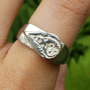 May include: A silver ring with a textured, triangular design. The ring is worn on a person's finger.