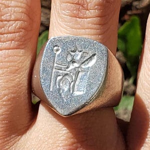 May include: A silver signet ring with a shield-shaped face. The face features a detailed embossed design of a crown, a staff, and other decorative elements. The ring is worn on a finger.