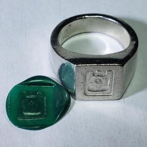 Może przedstawiać: Srebrny sygnet z kwadratową tarczą, obok zielonej pieczęci woskowej. Pierścień ma prostokątną tarczę z szczegółowym wzorem. Zielona pieczęć woskowa ma pasujący wzór, prawdopodobnie do odbijania.