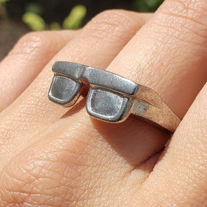 May include: A silver ring shaped like a pair of sunglasses. The ring is worn on a person's finger.