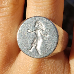 May include: A silver-colored signet ring featuring a detailed embossed image of a woman. The ring has a round face with a raised image of a woman with flowing hair and outstretched arms. The ring is worn on a finger.