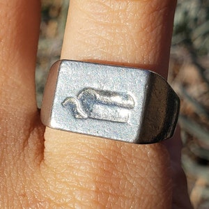 Könnte beinhalten: Ein silberner Siegelring mit einer rechteckigen Fläche, die ein stilisiertes Design einer Kreatur zeigt. Der Ring wird an einem Finger getragen.