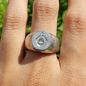 Pode incluir: Um anel de prata com um design circular que apresenta um símbolo em relevo. O anel é usado no dedo médio de uma mão.