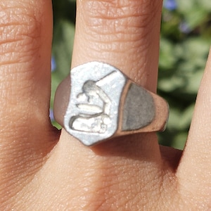 May include: A silver signet ring with a raised design of a person kneeling. The ring is worn on a person's finger.