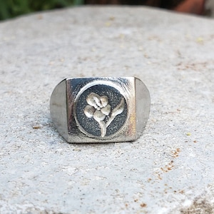 May include: A silver signet ring with a square face featuring a floral design.