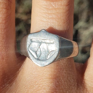 May include: A silver ring with a shield-shaped design. The shield has a raised design of two overlapping circles.