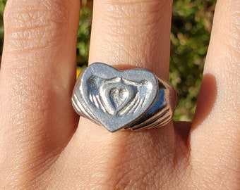 May include: A silver ring with a heart-shaped design featuring two hands holding a heart. The ring is worn on a person's finger.