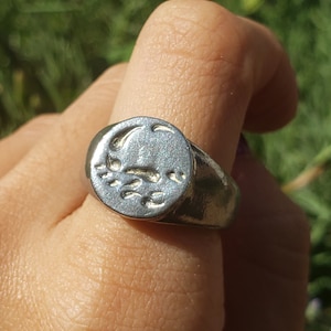 May include: A silver ring with a raised, circular design featuring a crescent moon and stars. The ring is worn on a finger.