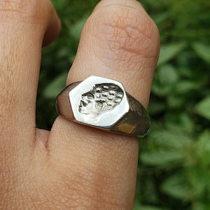 Puede incluir: Un anillo de sello de plata con un diseño detallado de un rostro humano. El anillo se lleva en un dedo.
