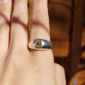 May include: A silver ring with a hexagonal, orange gemstone set in the center. The ring is worn on the middle finger of a hand.