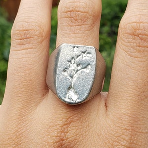 以下が含まれることがあります： 木製のデザインが施された銀の指輪。指輪は人の手の真ん中の指につけられています。