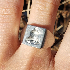 Può includere: Anello color argento con una superficie rettangolare con un design dettagliato a campana. L'anello ha una fascia liscia e un'emblema a campana strutturato. L'anello è mostrato su un dito. Il design è semplice.