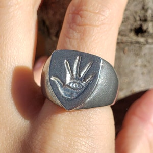 May include: A silver-colored ring with a shield-shaped face. The ring features a hand with an eye in the palm. The ring is worn on a finger.